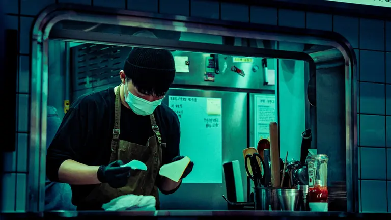 Masked chef working in an industrial kitchen, focused on food preparation.
