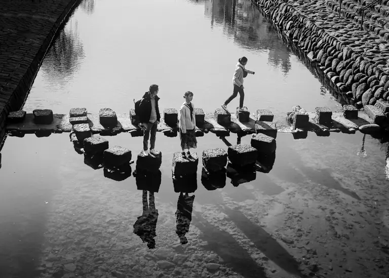 Three people crossing a stone path over water in Nagasaki, Japan, with clear reflections.